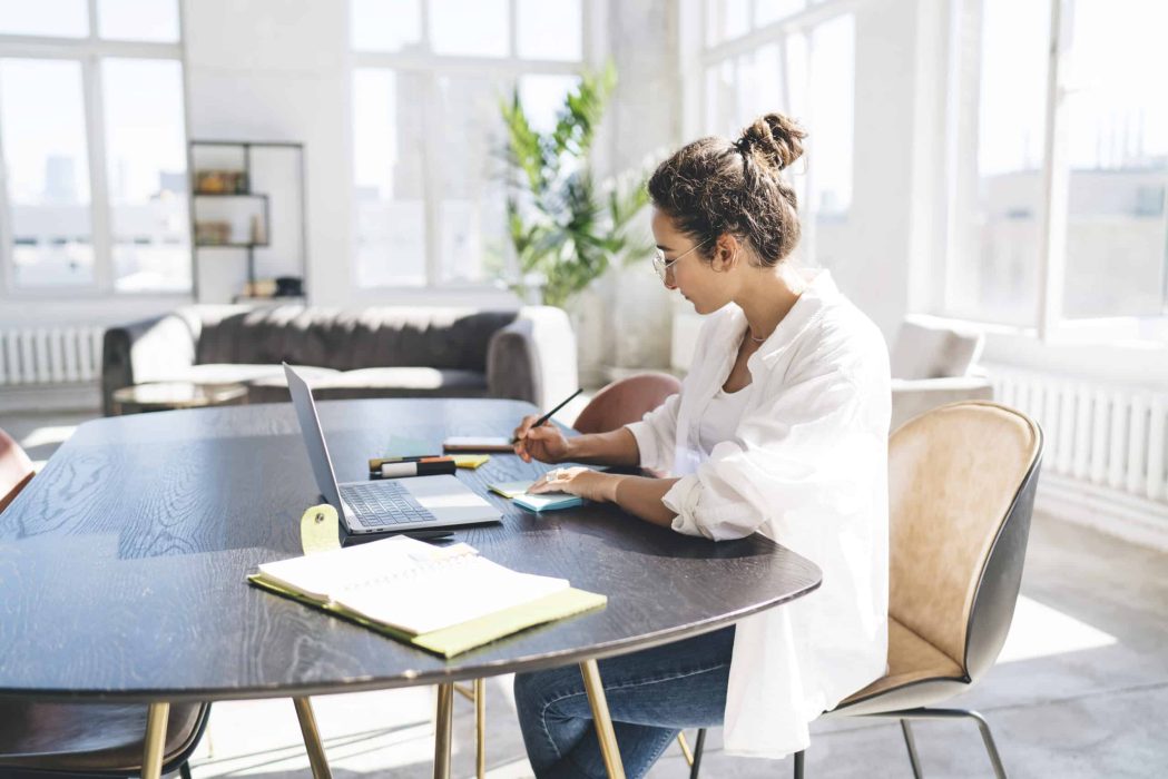 Side view of busy dark haired woman sitting at table with laptop and writing on sticky notes while working on business startup in living room at home